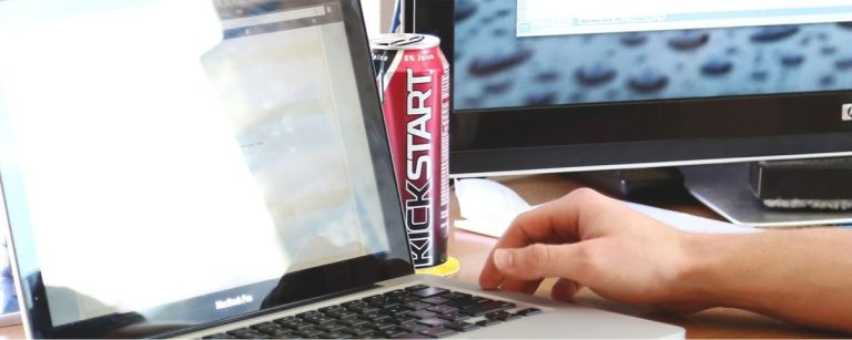 Ein Mann tippt auf seinem Laptop, neben ihm eine Kickstart-Dose und ein HP-Monitor mit Regentropfen.