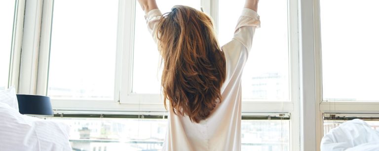 Frau streckt sich im Bett vor einem hellen Fenster, bereit für einen energiegeladenen Tag – perfekt für Fitness- und Ernährungsprogramme.