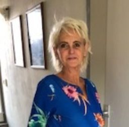 Frau mit kurzen blonden Haaren und einem blauen Kleid mit Blumenmuster lächelt in die Kamera.