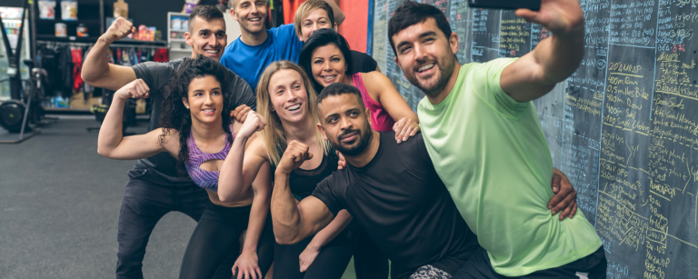 Nieuwsbrief juiste formaat (1) Gruppe von glücklichen Fitness-Enthusiasten posiert für ein Selfie im Fitnessstudio vor einer Tafel mit Trainingsübungen und Notizen.